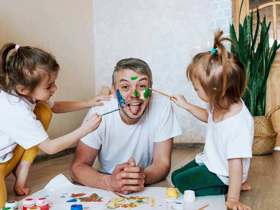 Ein männlicher Erzieher lässt sich in der Kita von zwei Kindern im Gesicht anmalen.