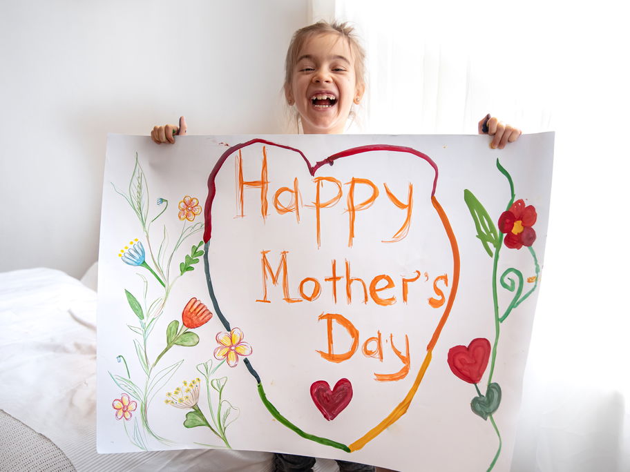 Ein kleines Mädchen hält ein selbst gebasteltes Bild mit der Aufschrift "Happy Mothers Day".