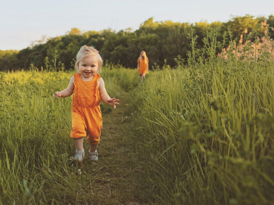 Ein kleines Mädchen in orangenem Sommeroutfit läuft durch eine Wiese.
