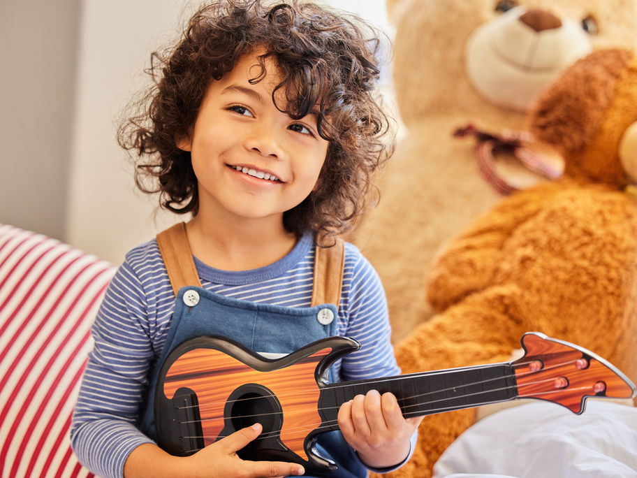 Ein Junge mit dunklen Locken sitzt mit seiner Gitarre im Kinderzimmer.