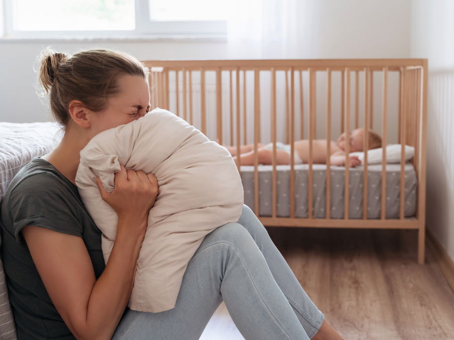 Eine Frau schreit in ein Kissen, während ihr Baby im Bett schläft