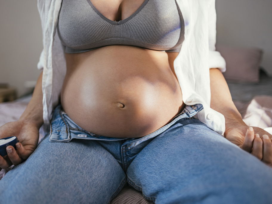 Eine Frau mit Babybauch sitzt mit geöffneter Hose auf dem Bett
