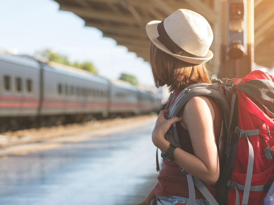 Ein junges Mädchen steht mit einem Rucksack an einem Bahngleis und wartet.