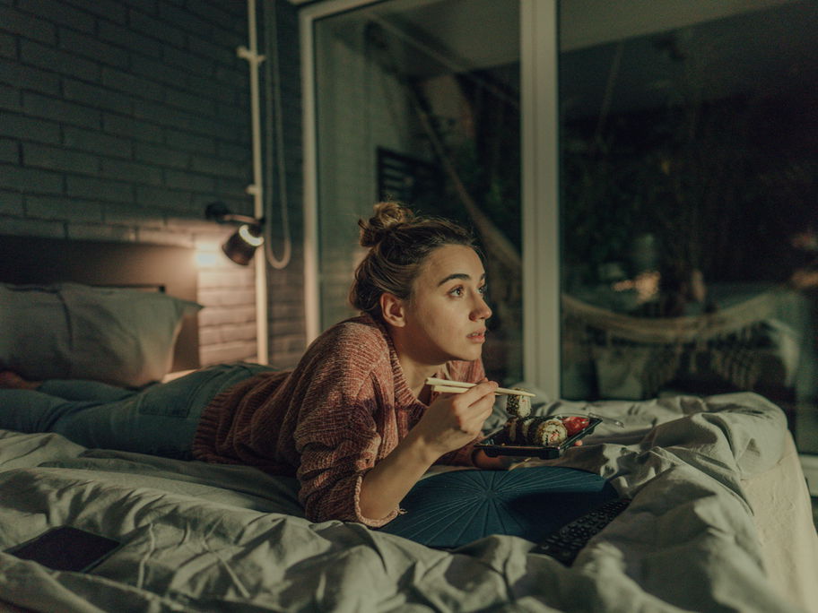 Eine Frau liegt abends im Bett, schaut TV und isst Sushi