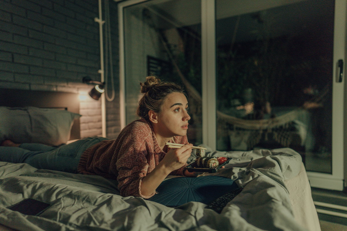 Eine Frau liegt abends im Bett, schaut TV und isst Sushi