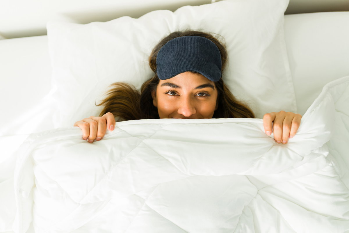 Eine Frau liegt mit Schlafbrille im Bett und zieht sich die Decke übers Gesicht.