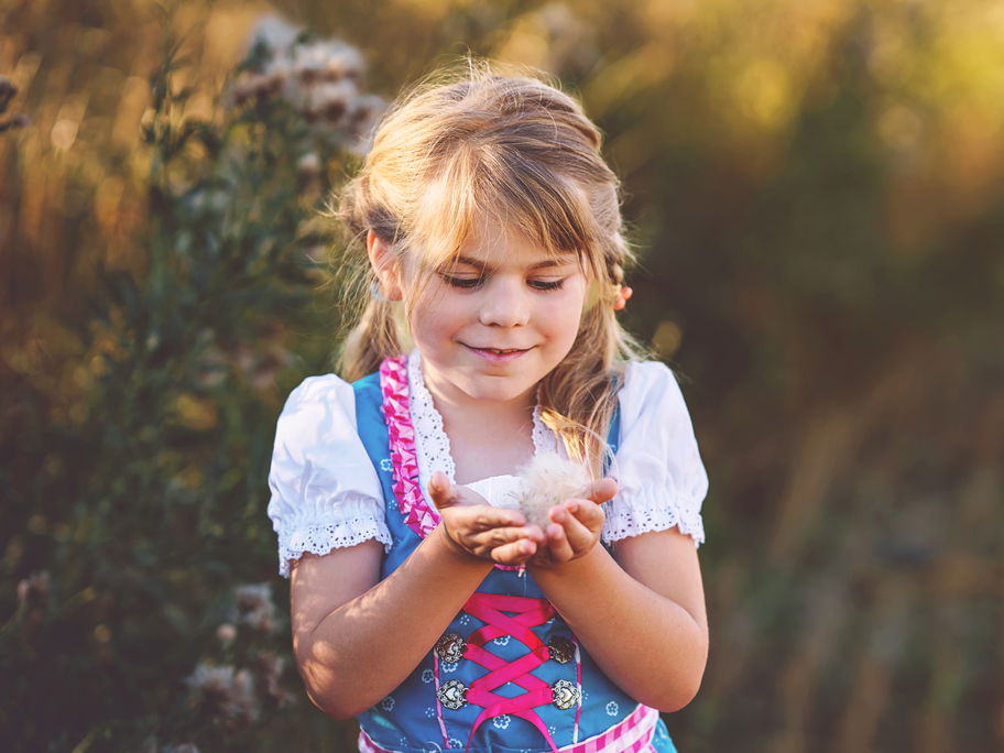 Ein kleines Mädchen im Dirndl steht auf dem Feld und hält eine Blüte in der Hand.