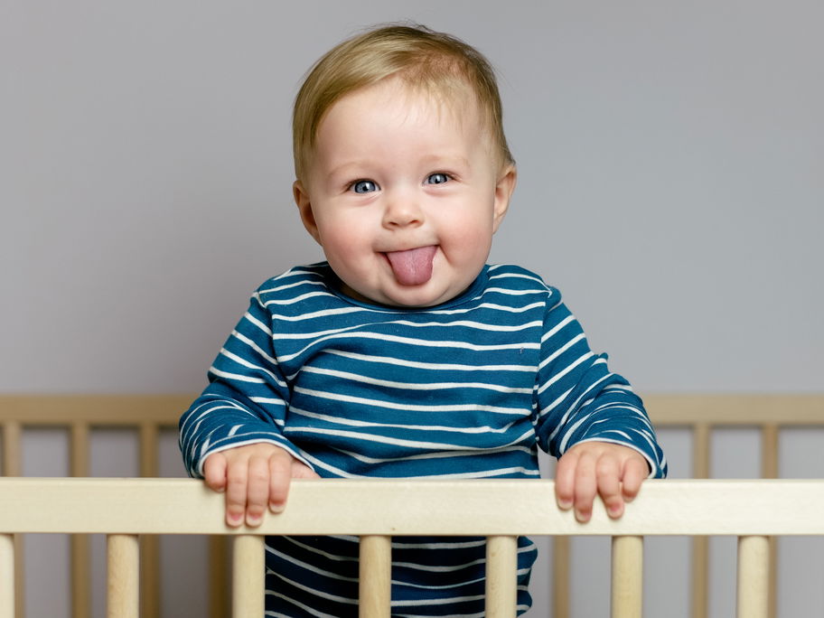 Ein kleiner Junge steht in seinem Gitterbett und streckt die Zunge raus.