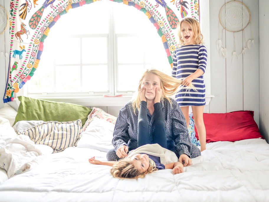 Ausgelaugte Mutter sitzt auf Bett, Kinder ziehen an ihren Haaren und machen Quatsch.