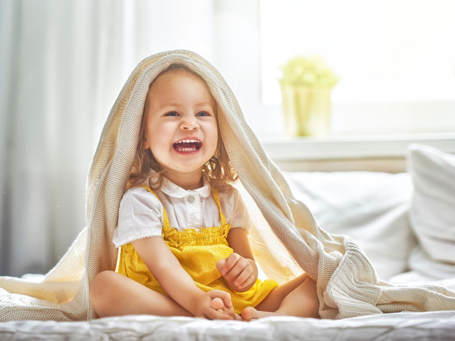 Ein kleines Mädchen sitzt lachend auf dem Bett und hat sich die Bettdecke über den Kopf geworfen.