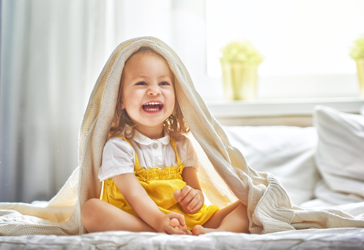 Ein kleines Mädchen sitzt lachend auf dem Bett und hat sich die Bettdecke über den Kopf geworfen.