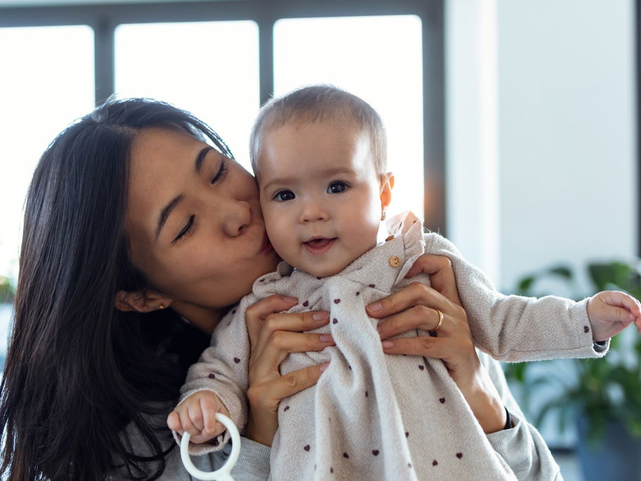 Eine asiatische Mutter hält ihr Baby-Mädchen hoch und küsst es zärtlich auf die Wange.