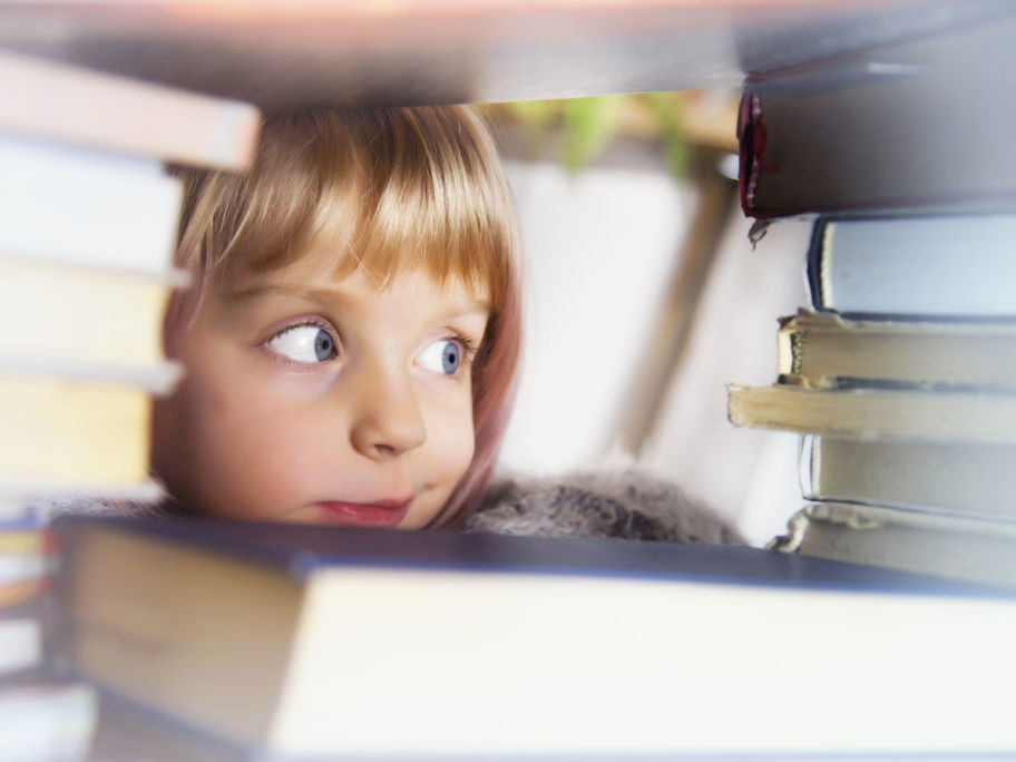 Mädchen schaut mit großen Augen zwischen Bücherstapeln hindurch