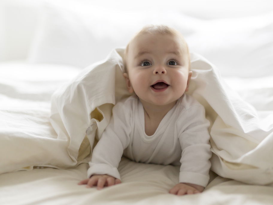 Ein kleiner Baby-Junge guckt frech unter einer weißen Bettdecke hervor.