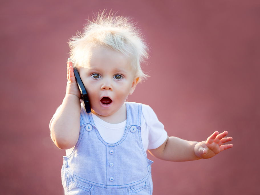 Ein kleiner Baby-Junge hält ein Handy in der Hand und schaut schockiert.