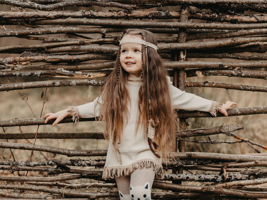 Ein kleines Baby-Mädchen im Boho-Look lehnt lässig an einem Holzzaun.