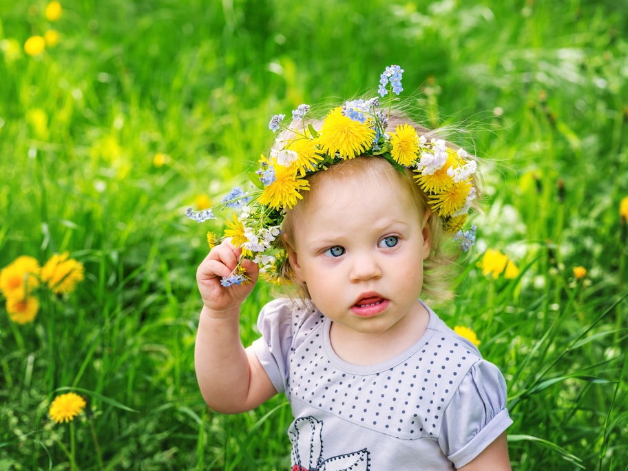 Kleines Mädchen mit Blumenkranz auf dem Haar steht auf einer Wiese.