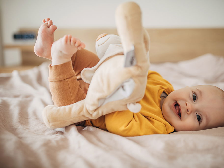 Kleiner Baby-Junge lacht fröhlich in die Kamera und hält ein Stofftier in den Händen.