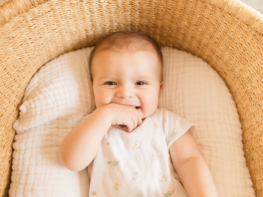 Ein kleiner, süßer Junge, der seine Finger im Mund hat und vom Babybett in die Kamera grinst.