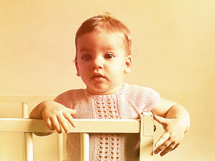 Ein Baby-Mädchen in den 60ern steht niedlich im Gitterbett