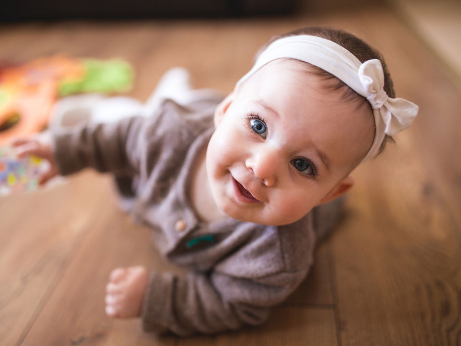 Kleines Baby-Mädchen mit Haarband liegt auf dem Boden und grinst.