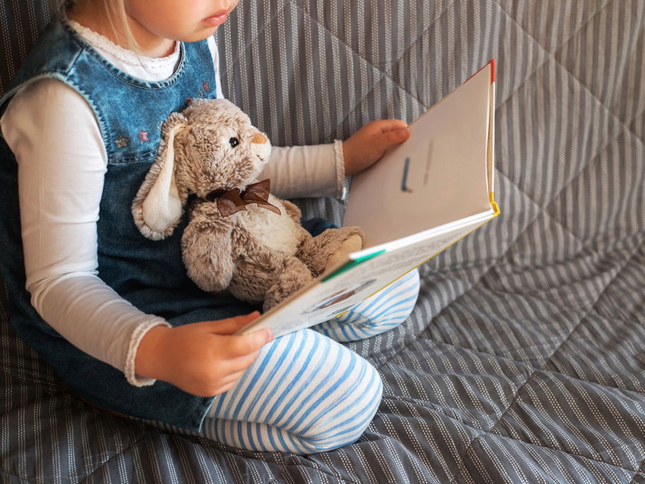 Kleines Mädchen sitzt auf einem Sofa, ein Hasen-Kuscheltier auf ihrem Schoß, und hält ein aufgeschlagenes Buch in der Hand.