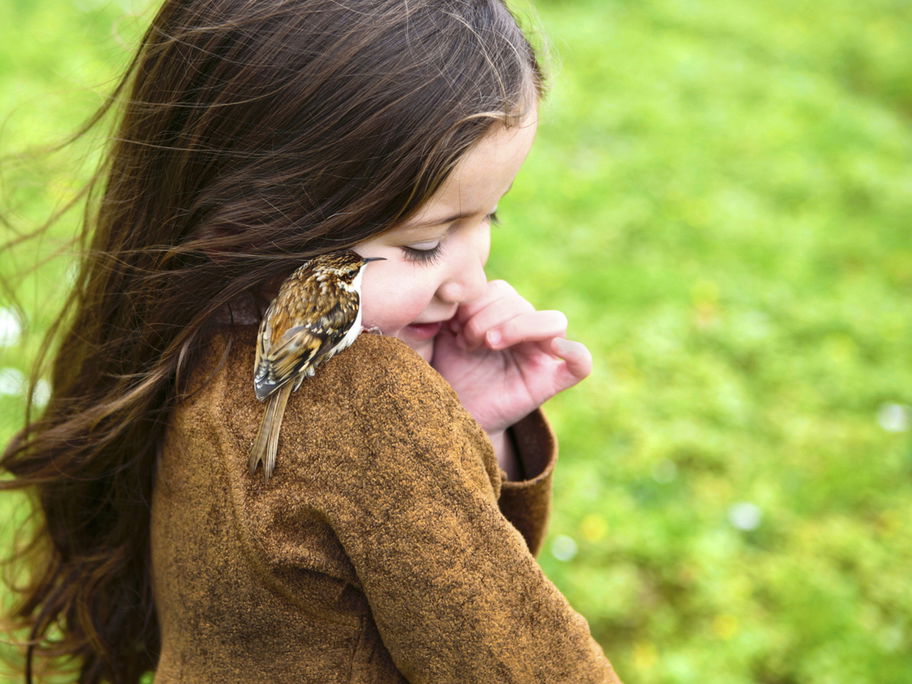 Ein Mädchen mit einem kleinen Vogel auf der Schulter steht auf einer Wiese.