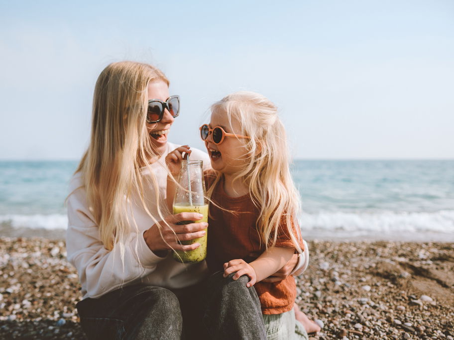 Mutter und Tochter sitzen fröhlich am schwedischen Strand, trinken einen Smoothie und lachen.