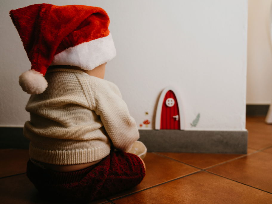 Baby-Junge mit Weihnachtsmütze sitzt vor der weihnachtlichen Wichteltür