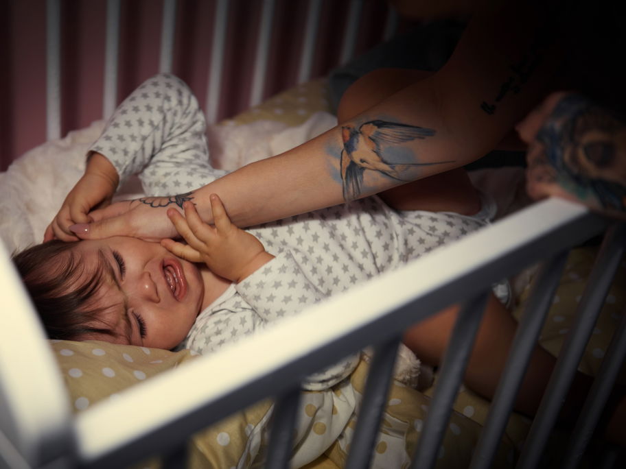 Ein weinendes Baby nachts im Babybett wird von Mutter gestreichelt.