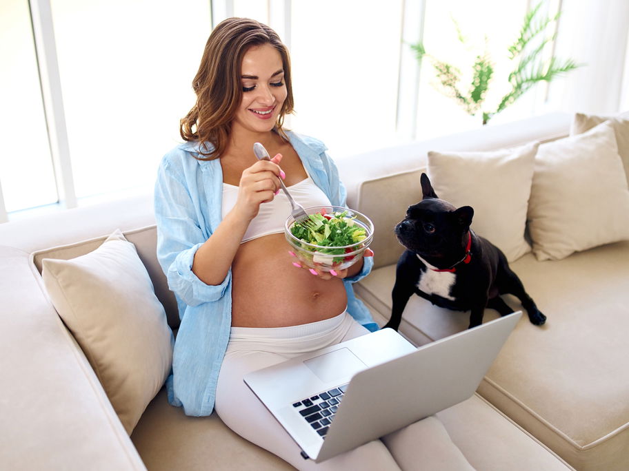 Schwangere sitzt auf Sofa und isst Salat.
