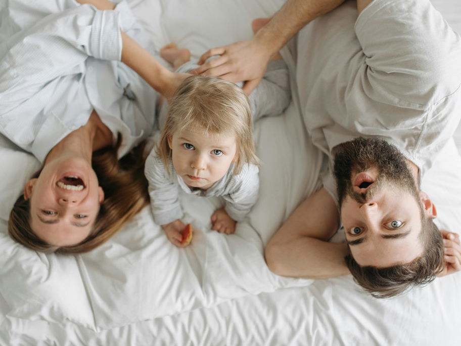 Eine glückliche Mutter liegt mit ihrem eher traurigen Kleinkind und neuen Partner im Bett.