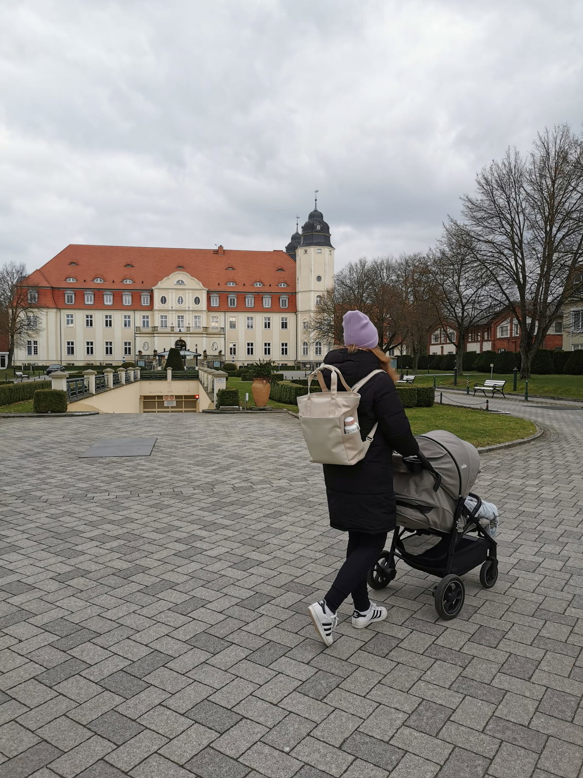 Eine junge Mutter läuft mit ihrem Kapten & Son-Wickelrucksack und Buggy vor einem Schloss.