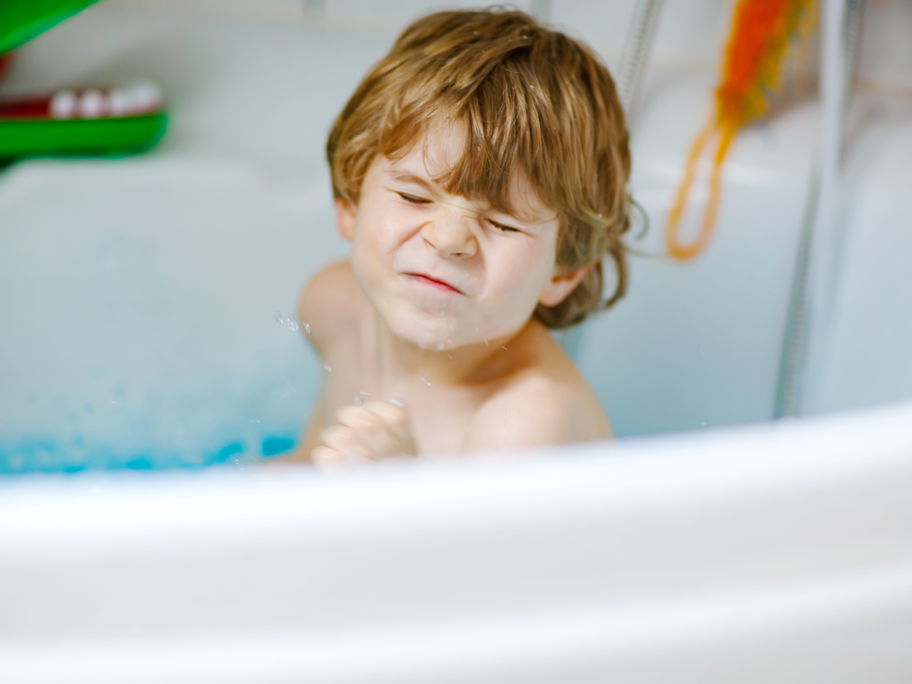 Ein kleiner Junge sitzt in der Badewanne und kneift die Augen zusammen.
