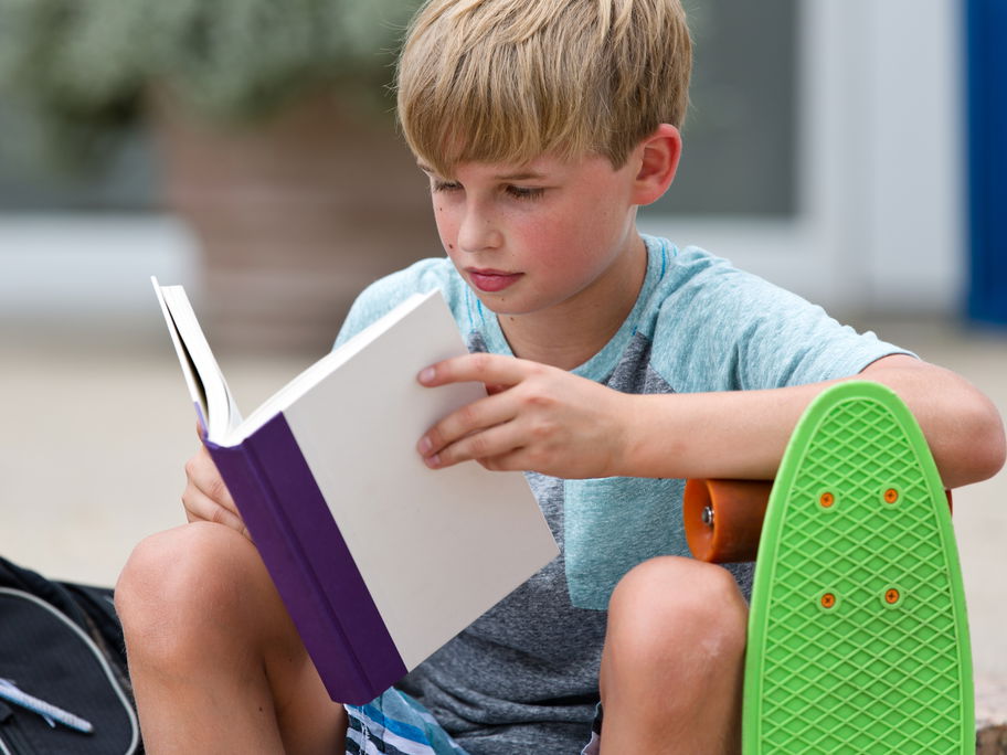 Junge mit Skateboard liest in einem Buch.