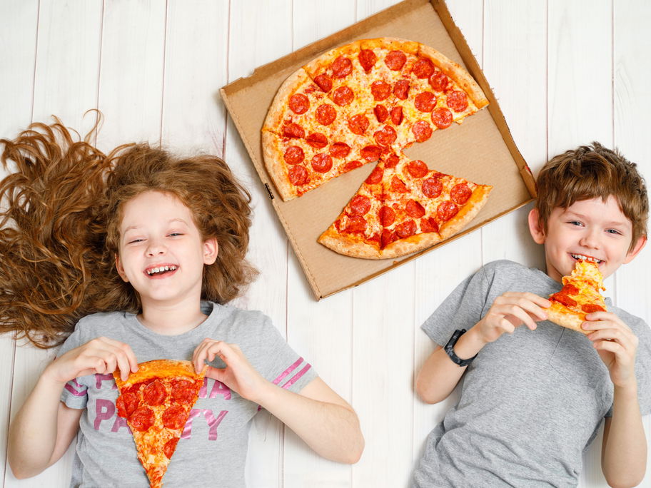 Zwei fröhliche Kinder liegen mit Salami-Pizza auf dem Boden.
