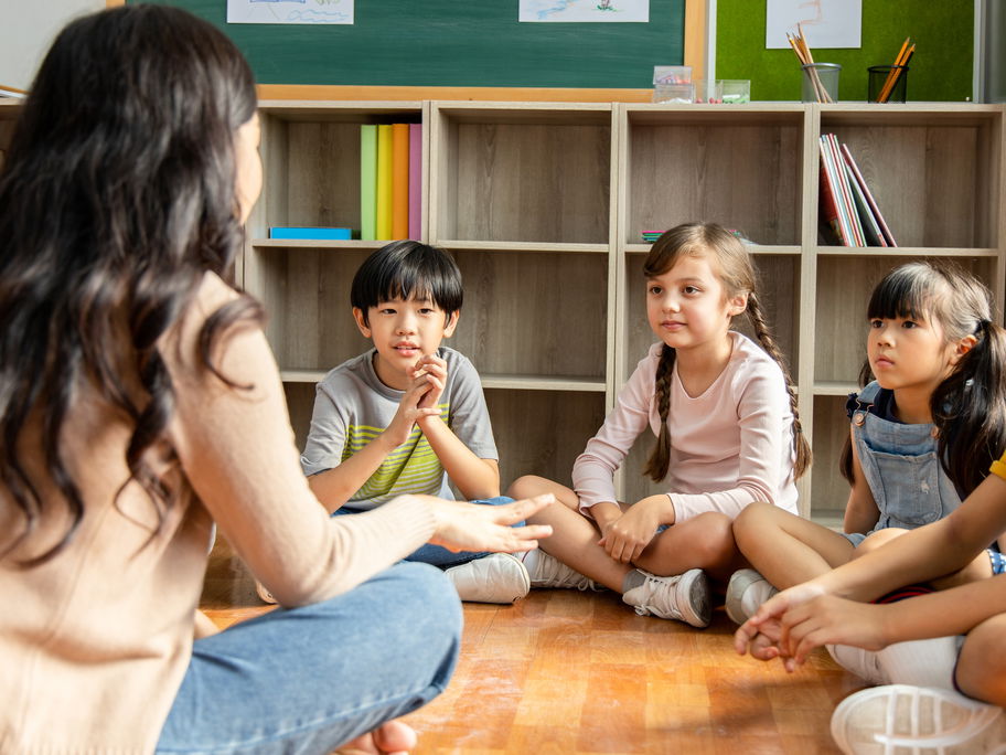 Lehrerin sitzt mit Grundschulkindern auf dem Boden.