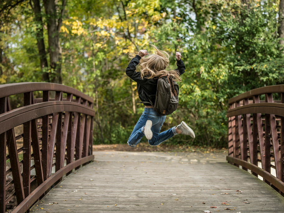 Mädchen mit Schulranzen springt hoch auf einer Holzbrücke.