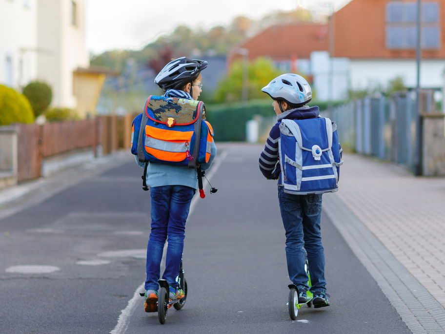 Zwei Kinder mit Schulranzen auf dem Rücken fahren auf Rollern zur Schule.