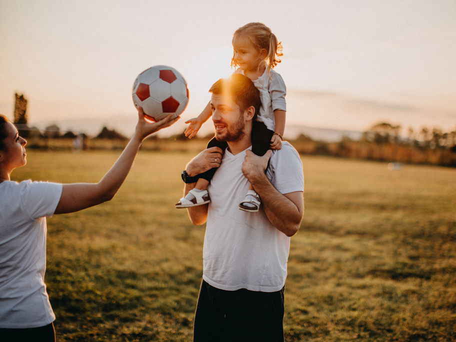 Mutter hält Mädchen einen Fußball hin, die auf Papas Schultern sitzt.