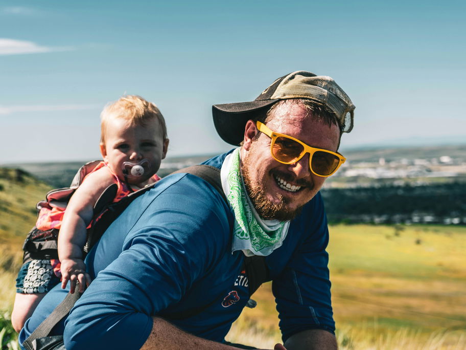 Fröhlicher Vater trägt Baby auf dem Rücken durch die Landschaft.