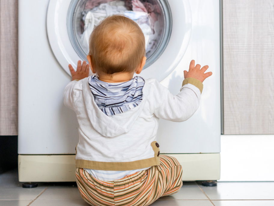 Baby sitzt vor der Waschmaschine und schaut hinein.