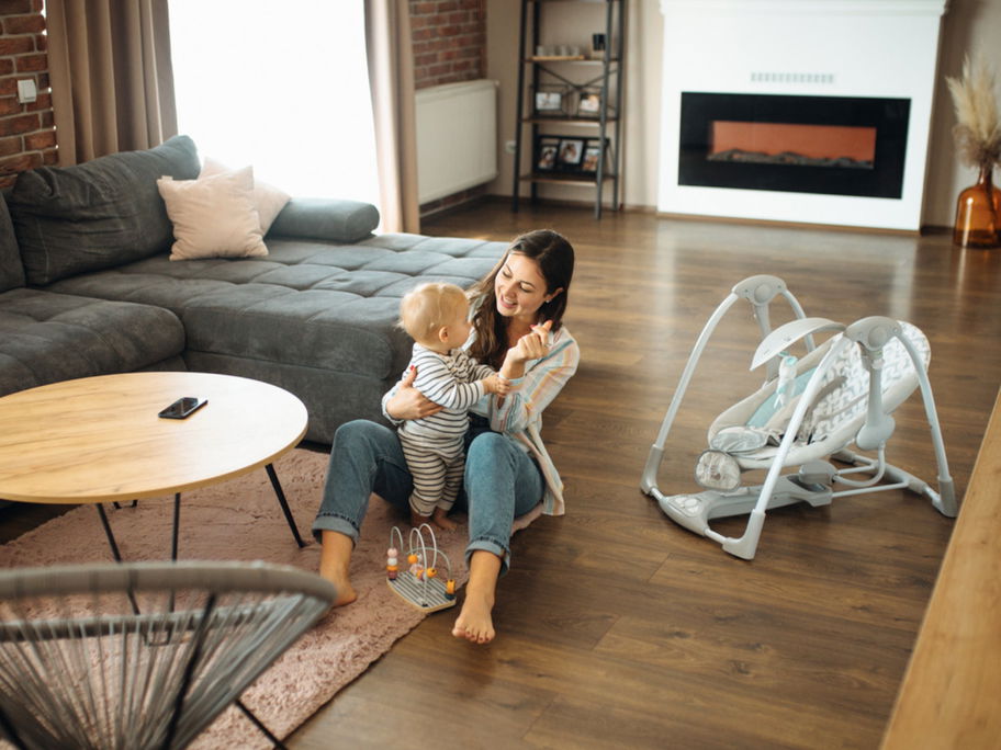 Mutter sitzt mit Baby auf dem Boden neben einer Babywippe.