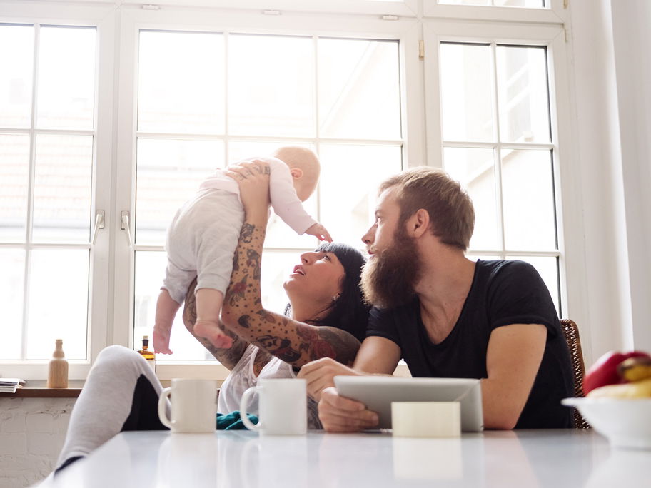 Eine Mutter hält ihr Baby in die Luft, der Vater sitzt daneben und schaut zu.