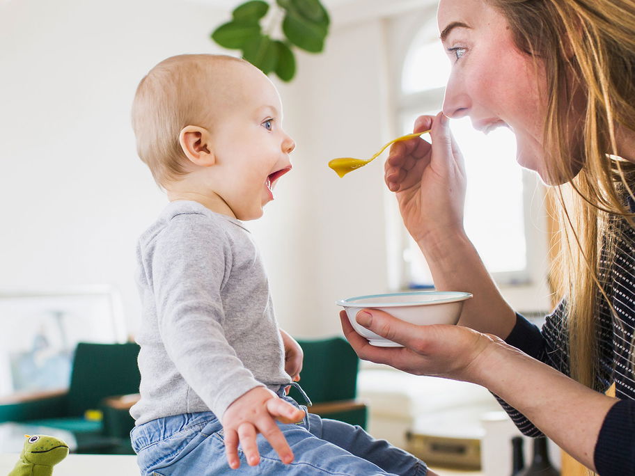 Mama füttert Baby mit Brei. Beide sperren den Mund auf.