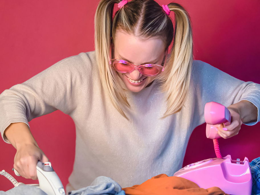 Fröhliche Frau mit pinker Brille bügelt und telefoniert gleichzeitig.