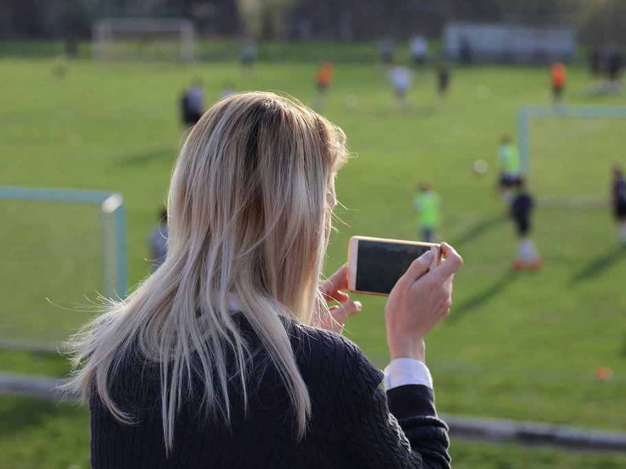 Mutter macht mit dem Smartphone Fotos vom Fußballspiel des Nachwuchses.