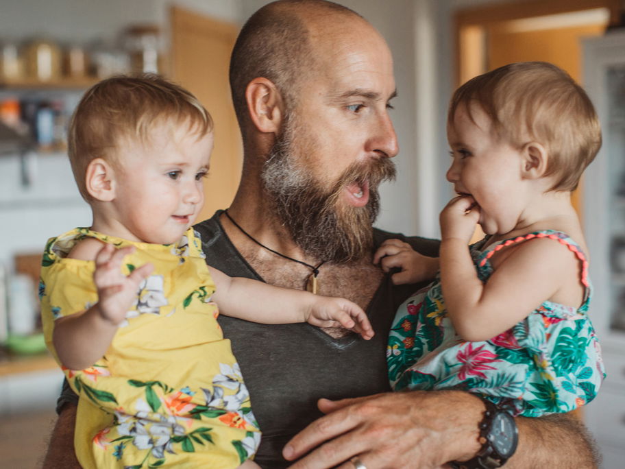 Papa mit Vollbart hat zwei Kinder auf dem Arm.