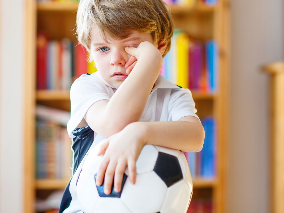 Krankes Kind reibt sich die Augen mit Fußball im Arm.